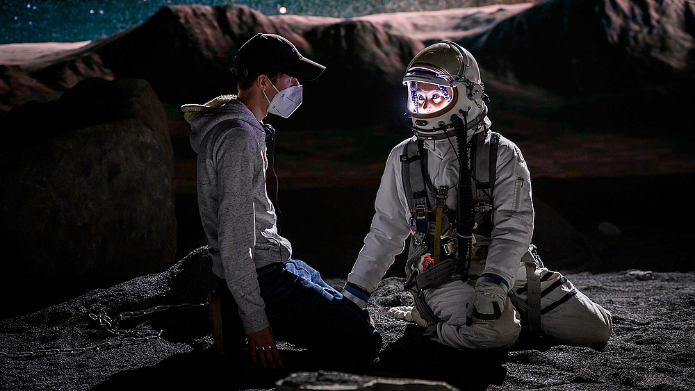 Eine männliche Person mit FFP2-Maske und Basecap sprihct mit einer weiblichen Person in einem Raumanzug. Beide knien in eine Mondlandschaft.