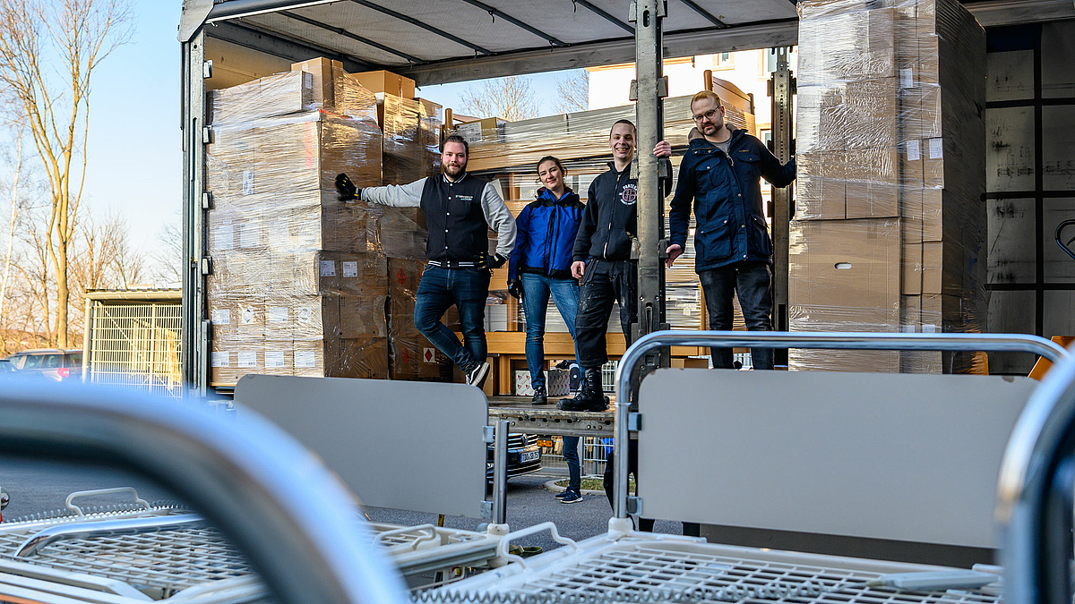 A young woman and three young men in work clothes are standing on a truck trailer whose side walls are open. Cartons that have already been loaded are stacked around them. In the foreground of the picture are still unloaded hospital beds.
