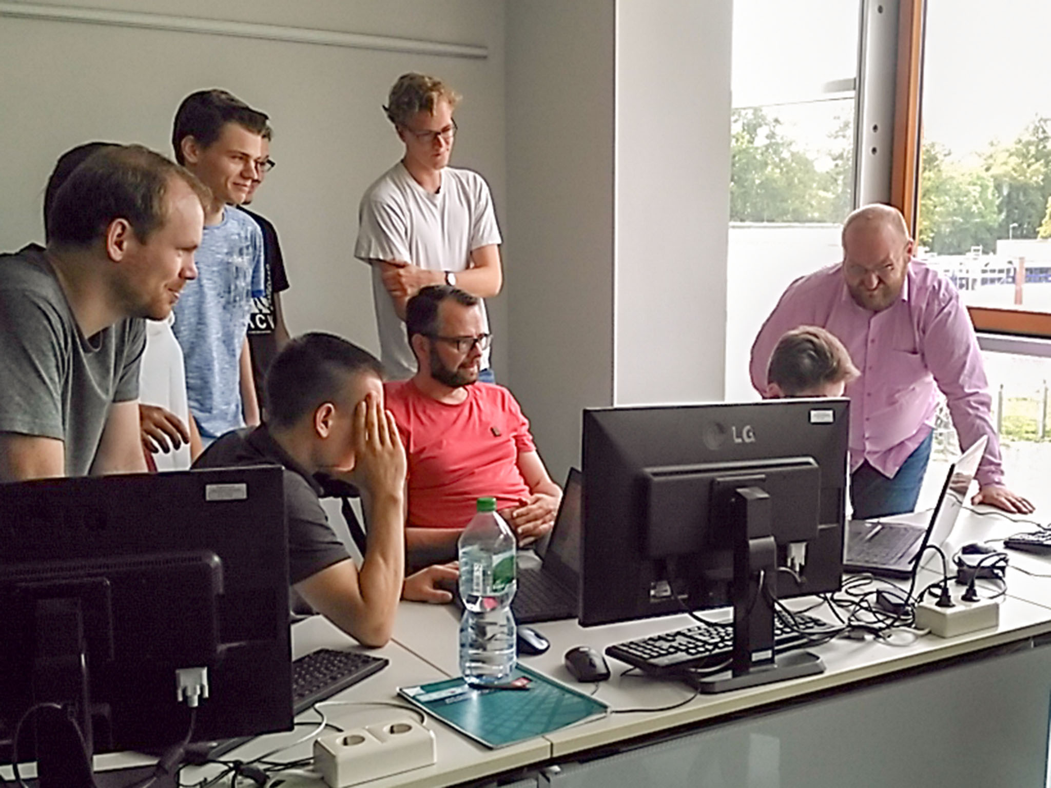 Bei der Summer School wurden Lösungsansätze für die selbstständige Gruppenarbeit zusammen diskutiert. Studierende und Dozenten stehen vor einem Schreibtisch mit Computer und diskutieren.