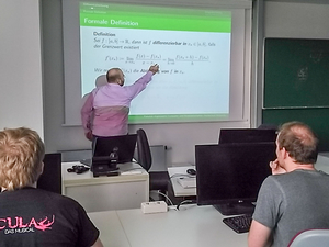 Dozent Erik Ludwig zeigt an einer Tafel auf eine mathematische Formel zur Definition der Differentiation, zwei Studierende hören ihm zu.