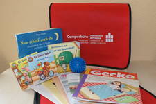 Beispielabbildung eines Willkommenspaketes rote Tasche im Hintergrund, davor einige Kinderbücher, ein blauer Massageball und Informationshefte