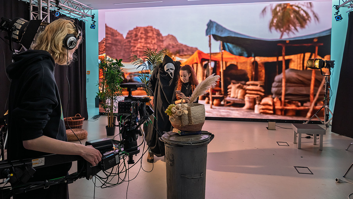 Blick am Kameramann vorbei auf eine Spielszene vor dem orientalischen Markthintergrund. In der Spielszene tritte eine Person mit schwarzen Umhang und Scary-Movie-Maske an eine junge Frau heran.