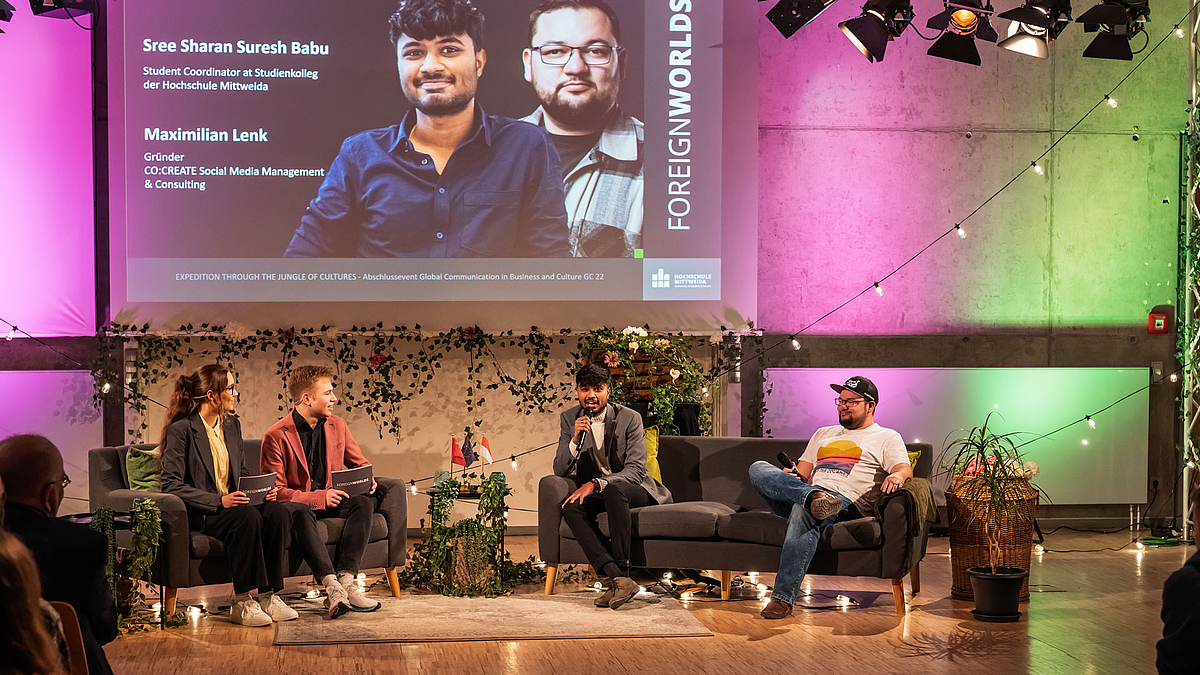 Auf dem Sofa neben den Moderator:innen sitzen zwei junge männliche Personen, von den eine in ein Handmikrofon spricht. Im Hintergrund sind die Porträts der beiden auf der Leinwand zu sehen sowie deren Namen und Tätigkeiten: „Sree Sharan Suresh Babu. Student Coordinator at Studienkolleg“ und „Maximilian Lenk. Gründer Co:Create Social Media Management & Consulting”.