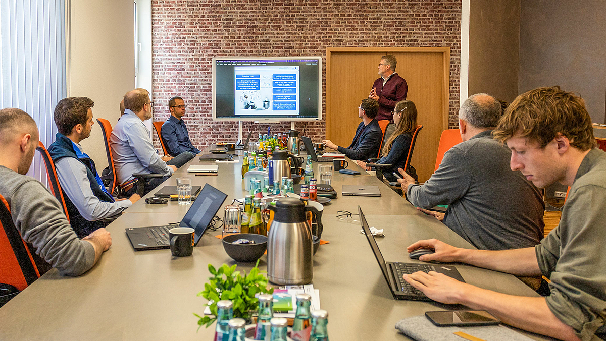 Das Foto zeigt 9 Personen an einem Konferenztisch. Eine davon ist verdeckt. Alle haben Laptops vor sich stehen. Rechts neben einem großen Monitor im Bildhintergrund steht eine weitere männliche Person. Der Inhalt der Präsentation auf dem Monitor ist nicht lesbar.