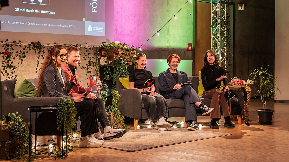 Die beiden Moderator:innen sitzen auf einem Sofa. Auf einem weiteren Sofa rechts von ihnen sitzen drei junge weibliche Personen.