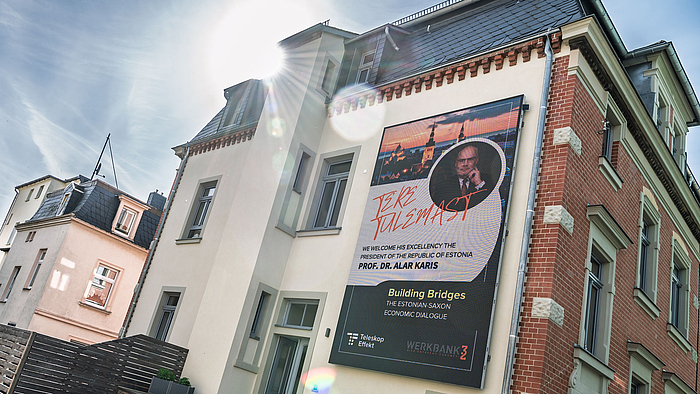 Eine Hausfassade mit einem wandfüllenden LED-Display: Es zeigt das Foto von Alar Karis und unter anderem den Text „Building Bridges“. Im gegen die Sonne gemachten Foto spiegeln sich die Linsen des Objektivs. 