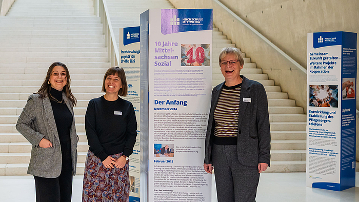 Sie sind die wissenschaftlichen Köpfe hinter Mittelsachsen Sozial: Professorin Isolde Heintze, Friederike Haubold und Tabea Esche (v.r.n.l.). Drei weibliche Personen fröhlich lachend. Hinter ihnen quaderförmige Aufsteller, darunter einer mit der Aufschrift „10 Jahre Mittelsachsen Sozial“ und weiterem Text.