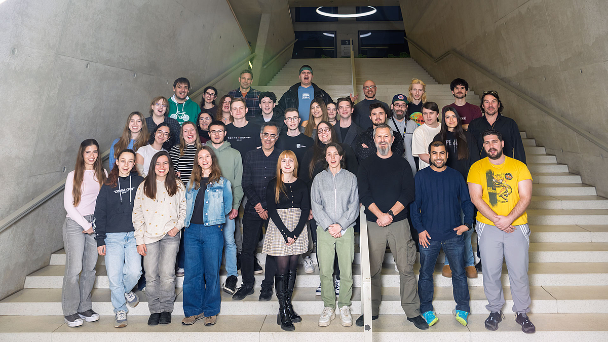 Etwa 35 meit junge Menschen stehen fröhlich lachend auf den ansteigenden Stufen einer großen Treppe im Inneren eines Gebäudes mit grauen Sichtbetonwänden.