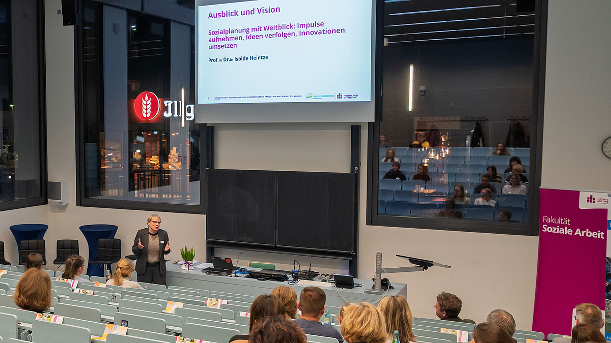 Abschluss mit Ausblick von Professorin Isolde Heintze. Weibliche Person spricht zum Publikum im Hördaal. Auf der Leiwnand über ihr ist zu lesen: "Ausblick und Vision Sozialplanung mit Weitblick: Impulse aufnehmen, Ideen verfolgen, Innovationen umsetzen."