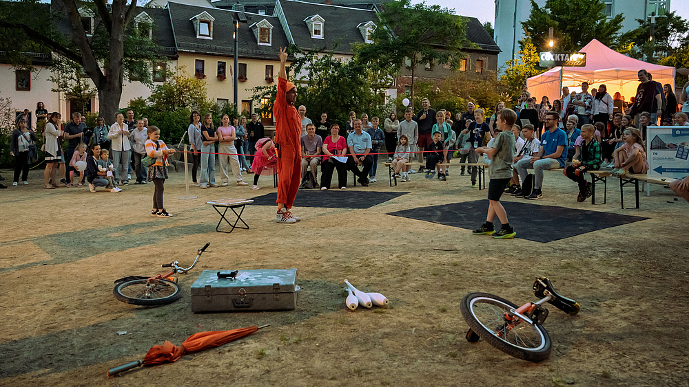 Ein Clown im orangenen Anzug steh in der Mitte zwichen zwei Kindern, die ein Gummiseil gespannt haben, das zwischen seinen Beinen entlang geführt ist. Im Hintegrund amüsieren sich zahlreiche Zuschauende unterschiedlichen Alters.