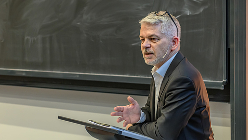 Eine männliche Person mit entschiedener Mine vor einer schlecht geputzten Tafel, die Brille nach oben ins Haar geschoben, den linken Ellbogen auf ein Rednerpult abgestützt und die rechte Hand geöffnet, spricht zu einem im Bild nicht sichtbaren Publikum. 