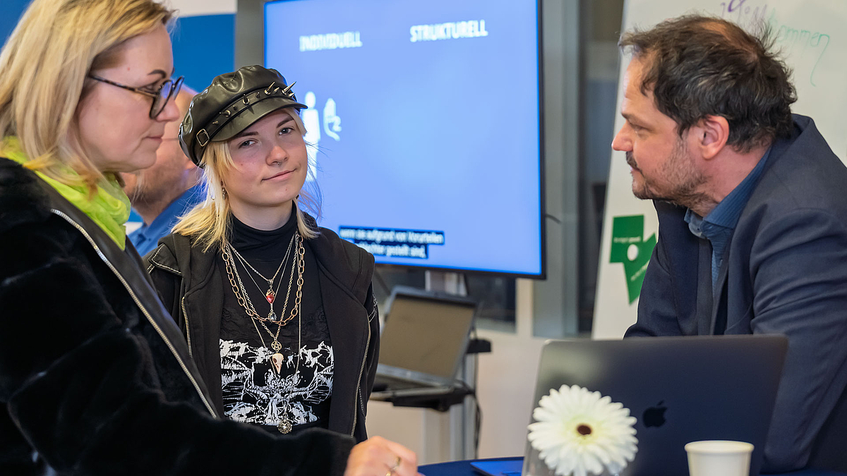 Zwei weibliche und eien männliche Person an einem Stehtisch im Gespräche. Die jüngere weibliche Person in der Mitte wendet im Gothic-Outfit mit schwarzer Matrosenmütze wendet ihren Blick der Kamera zu.