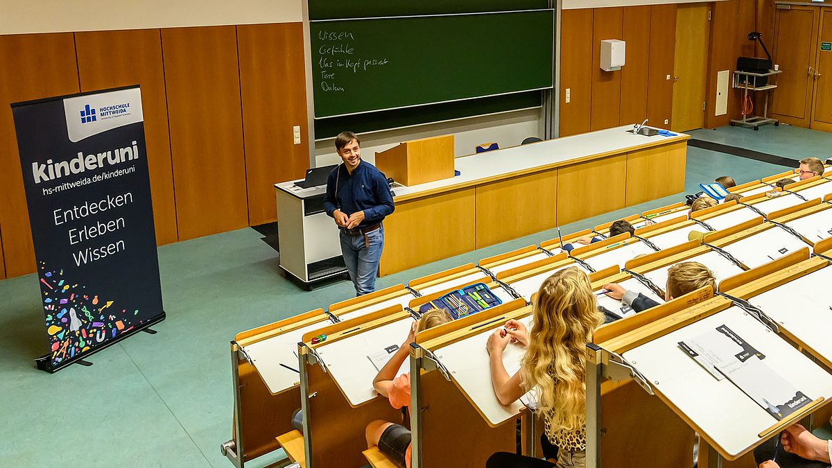 Dozent wendet sich freudnlich seinen jungen Zuhörer:innen zu. Links steht ein Rollup mit der Aufschrift: "Kinderuni. Entdecken, Erleben, Wissen"