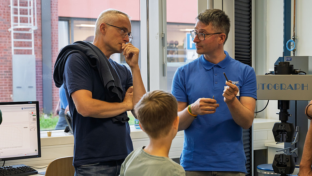 Zwei männliche Personen vor einen Festigkeitsprüfstand. Eine Person hält ein Werkstück in der Hand