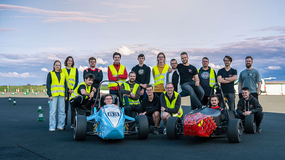 Gruppenfoto mit 18 jungen Personen, teilweise in gelben Warnwesten, und einem blauen und einem roten offenen Rennwagen auf einer geteerten Fläche. Zwei der Personen sitzen in den Rennwagen, fünf knien links und rechts davon, die anderen stehen dahinter. Im Bildhintergrund sind rot-weiße Pylone aufgestellt.