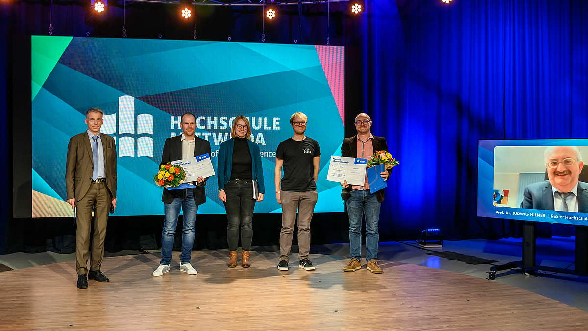 Das Foto zeigt fünf Personen auf einem Podest im Studio, darunter die Preisträger mit Blumen und Urkunden. Am rechten Bildrand ist ein Monitor mit einer weiteren zugschaltetet Person zu sehen, dem Rektor der Hochschule Mittweida.