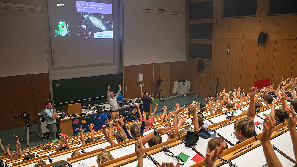 Großer Hörsaal der Kinderuni Mittweida, voll besetzt mit Kindern, die beide Hände hochhalten. Vorne stehen zwei Professoren, auf der Leinwand ist eine Galaxie und das Thema ‚Mit dem Licht auf Gullivers Reisen‘ zu sehen.