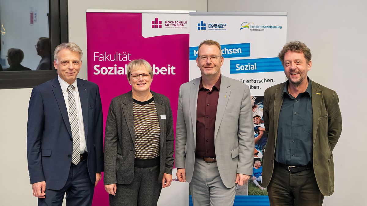 Rektor Volker Tolkmitt, Projektleiterin Isolde Heintze, Landrat Sven Krüger, Dekan Stephan Beetz (v.l.n.r.). Gruppenbild mit drei männlichen und einer weiblichen Person am unteren Endes des Hörsaals vor Rollups mit "Fakultät Soziale Arbeit" und "Mittelsachsen Sozial"."
