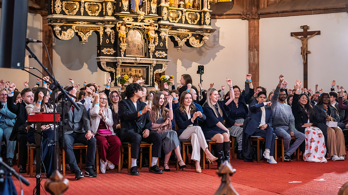 Junge Menschen sitzen auf Stühlen im Kirchenschiff und halten iher Arme nach oben. An den Handgelenken tragen sie Bändchen.