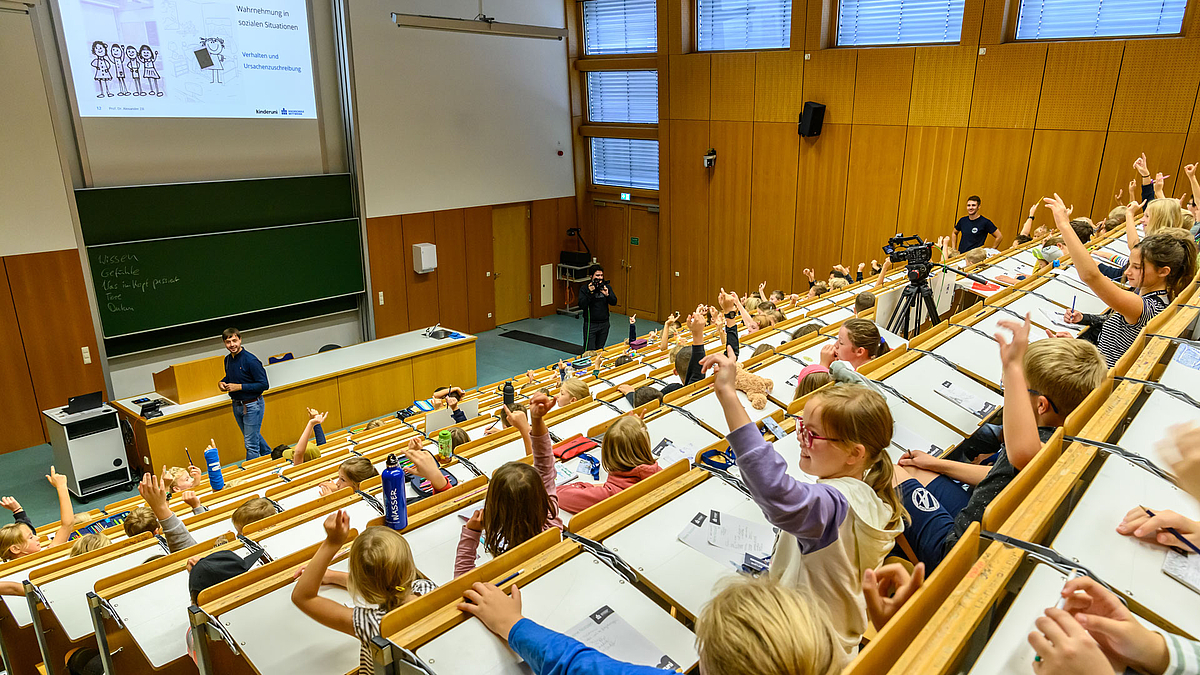 Viele sichmit ausgetreckten Armen und Fingern sich meldende Kinder in den aufsteigenden Bankreihen des Hörsaals.