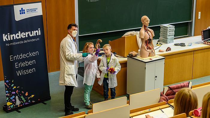 Neben dem Pult in einem Hörsaal stehen eine erwachsene männliche Person mit Maske sowie ein Mädchen und ein Junge, alle tagen weiße Laborkittel. Links seht ein Rollup mit der Aufschrift „kinderuni, Entdecken, Erleben, Wissen.“ Vor dem Pult steht auf einem Wagen ein lebensgroßes menschliches Torso -Anatomie-Modell mit herausnehmbaren Organen. Das Mädchen hält ein Stoff-Modell nach oben, das das Ebola-Virus darstellt. Der Junge hat die Stoff-Nachbildung des Coronavirus in der Hand.  