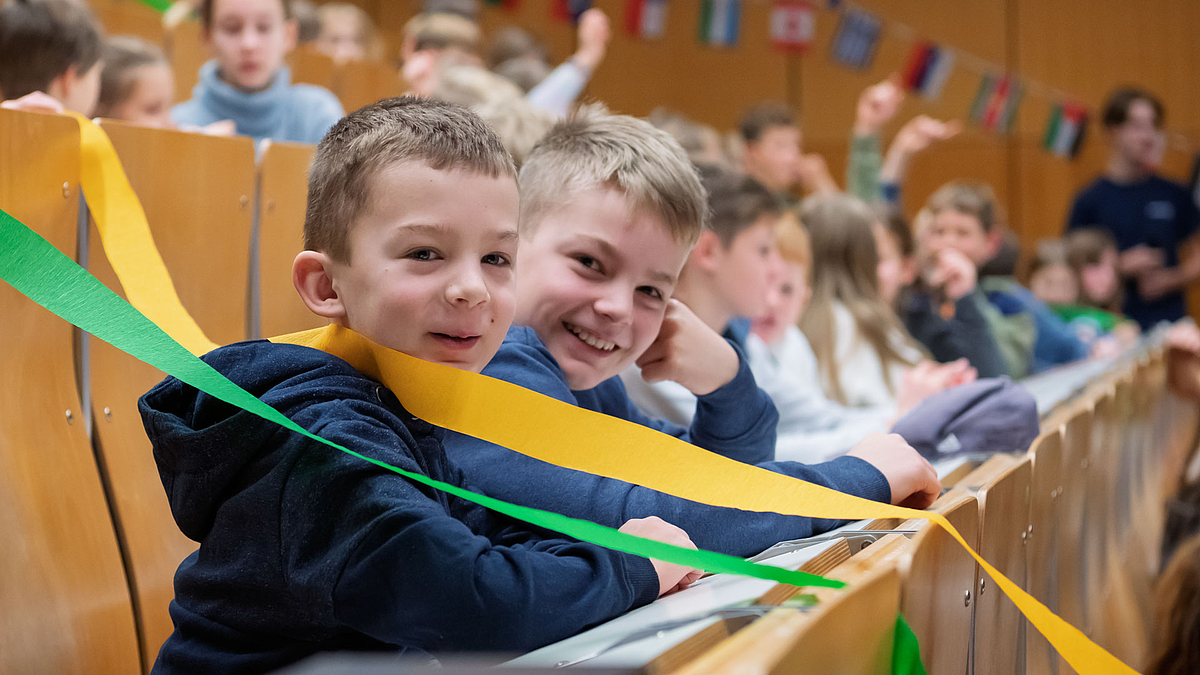 Kinder im Hörsaal spannen farbige Bände kreuz und quer über die Sitzreihen.