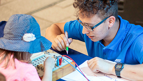 Kevin Blümel studiert Elektrotechnik und engagiert sich vielfältig an der Hochschule Mittweida. Kevin Blümel betreut bei der Nacht der Wissenschaften eine Elektrotechik-Station. Vor ihm sitzt ein junges Mädchen, das eine experimentelle Schaltung aufbaut.