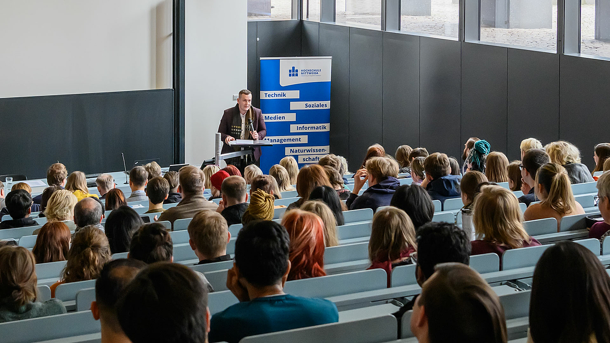 Blick von hinten in einen besetzten Hörsaal. Unten im Hörsaal im Bildhintergrund steht eine männliche Person an einem Pult, dahinter das Rollup mit dem Logo der Hochschule Mittweida und den Begriffen „Technik“, „Soziales“, „Medien“, Informatik“, Management“ und „Naturwissenschaft“.