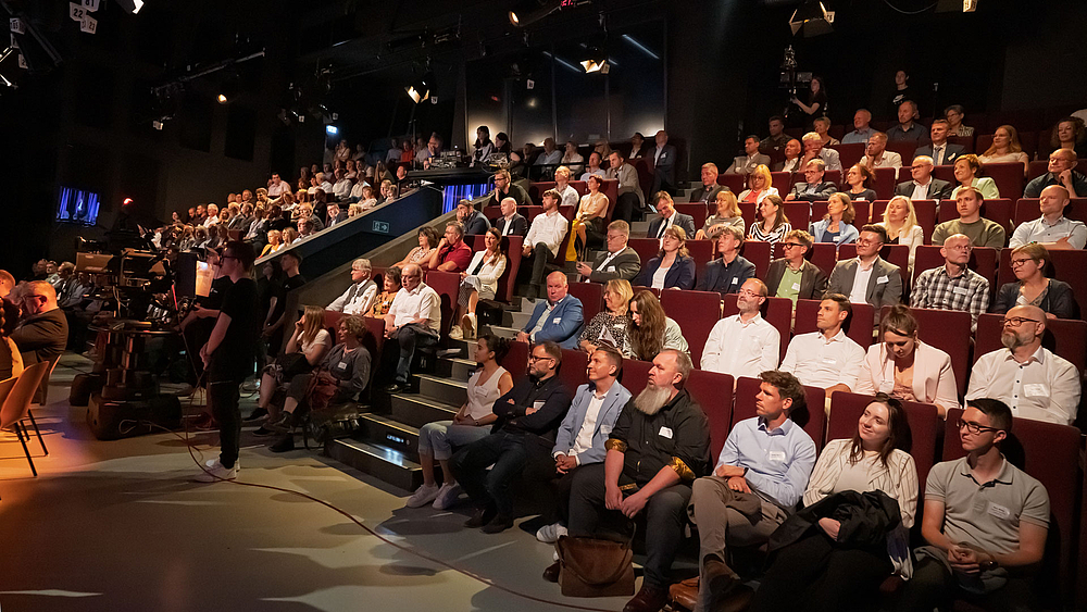 Volle Zuschauerränge im Fernsehstudio. Im Vordergrund sind von hinten zwei Musiker einer Band von hinten zu sehen.