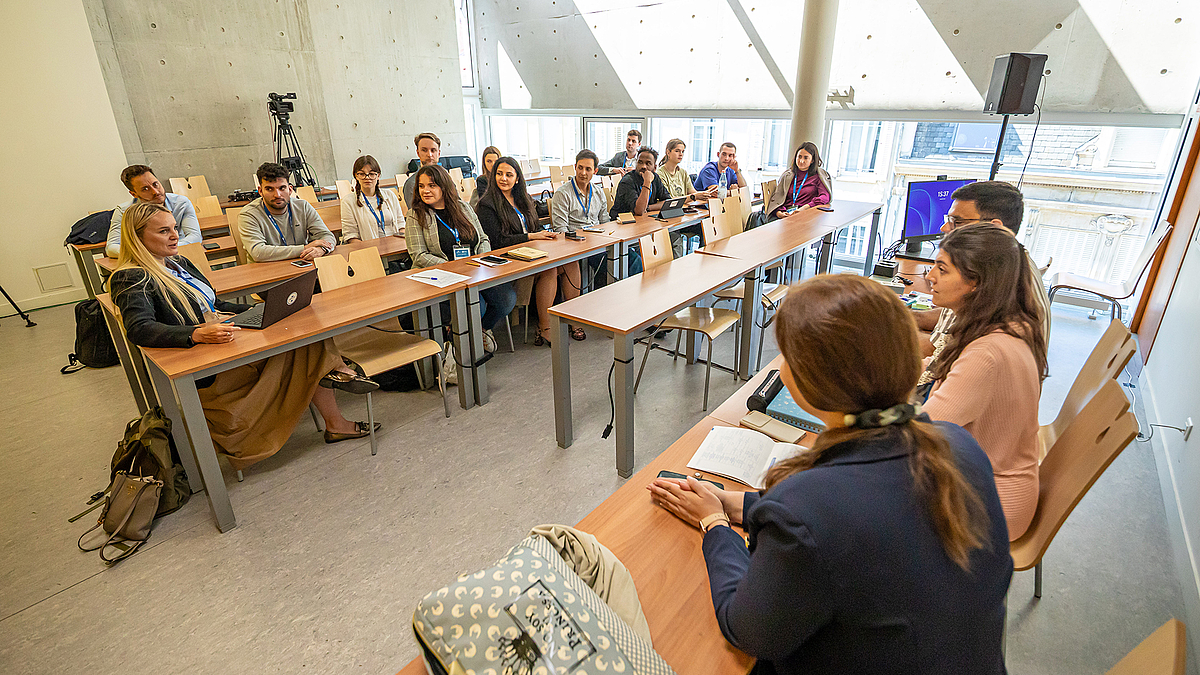 Eine Gruppe von über 20 Studierenden sitzt in einem Seminarraum. Drei sitzen vorne und schauen die große Gruppe an. In der ersten Reihe sitzt eine etwas ältere Frau und hört der sprechenden Vorsitzenden zu.