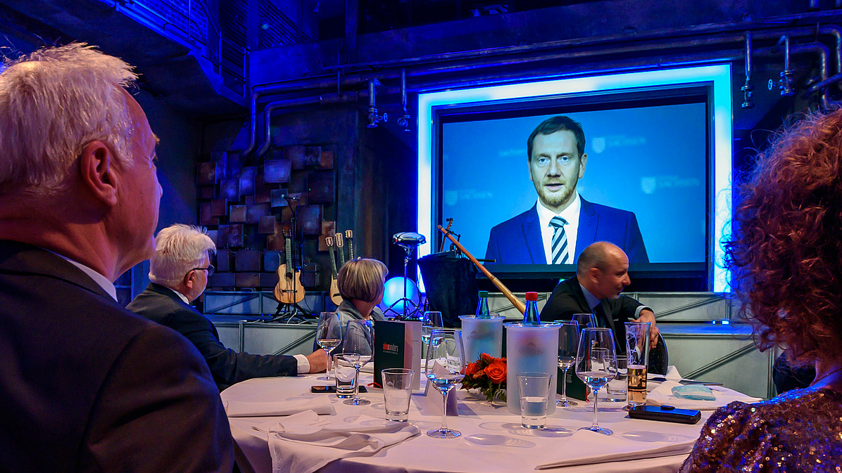 Das Foto zeigt im Vordergrund einen runden Tisch mit Personen von hinten fotografiert. Im Bildhintergrund auf der Leinwand ist der Sächsische Ministerpräsident Michael Kretschmer in seiner Videobotschaft zu sehen.