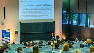 Blick über die Köpfe der Zuhörenden im Hörsaal hinweg auf die Dozentin vorne am Pult. Über ihr ist eine Folie projiziert der Überschrift „Was nehmen wir mit?“