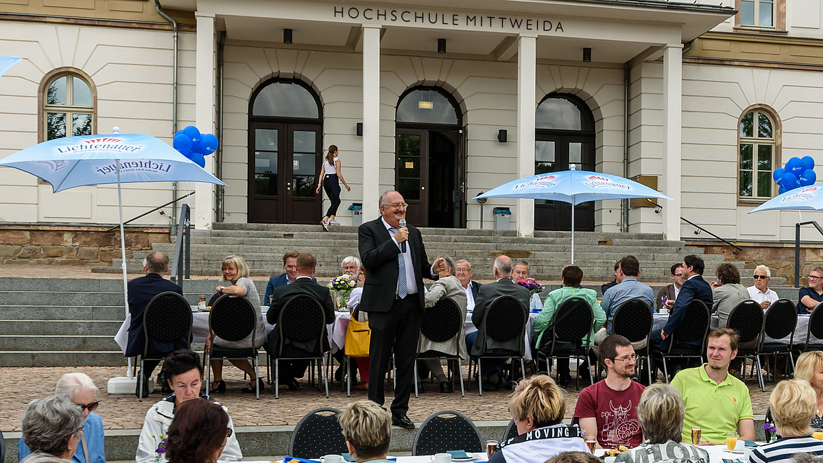 Das Foto zeigt eine männliche Person stehend auf einem Platz im Freien, vor und hinter der Person sitzen Menschen an Tischen. Im Bildhintergund ist das Treppenportal des Hauptgebäudes der Hochschule zu sehen mit der Aufschrift Hochschule Mittweida.