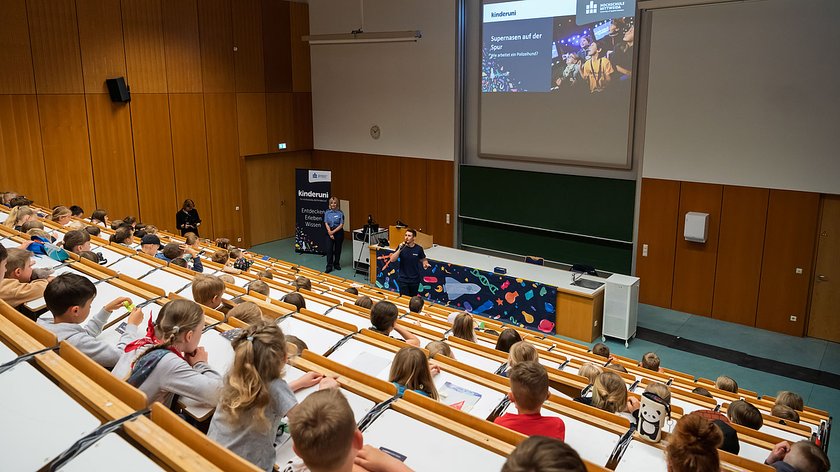 Vorlesungssaal mit Kindern, ein Moderator steht mit einem Mikrofon in der Hand vor dem Pult. Auf der der Leinwand hinter ihm ist die Folie mit dem Titel der Vorlesung projiziert: „Supernasen auf der Spur. Wie arbeitet ein Polizeihund?“