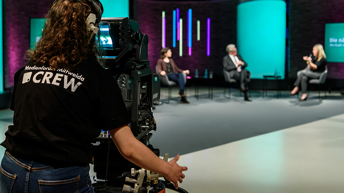 Das Bild zeigt eine Studentin an einer Studio-Fernsehkamera von hinten. Im Hintergrund sin unscharf drei Podiumsteilnehmende zu erkennen.