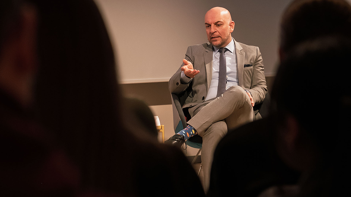 Blick zwichen den Köpfen von Zuschauenden auf männliche Person im Podium, die spricht und gestikuliert.
