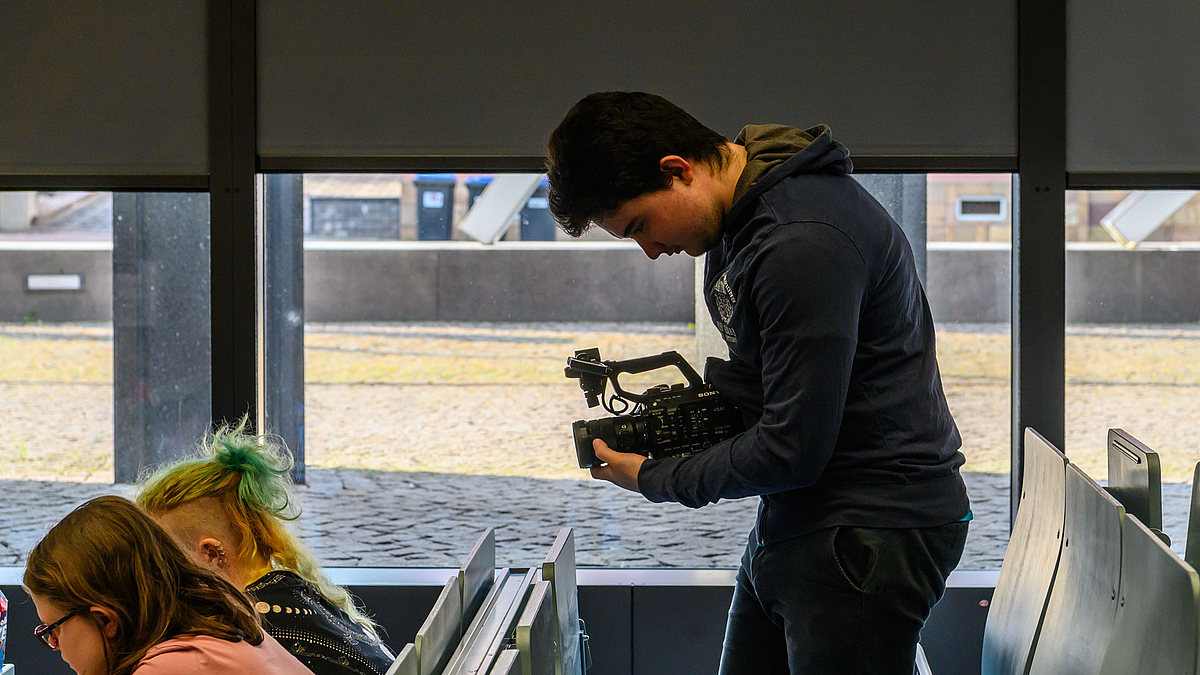 Ein junger Mann steht neben einer Bankreihe im Hörsaal, hält eine Videokamera in Bauchhöhe.