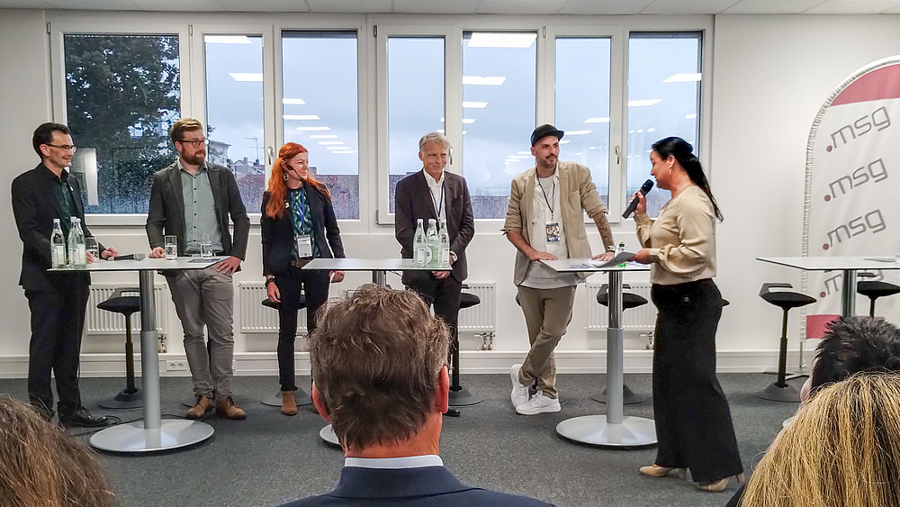 Podium in Passau: MIKOMI-Wirtschaftsdialog mit Rektor Gerd Strohmeier (TU Chemnitz), Dr. Oliver Georgi (VibroCut GmbH), Katrin Hoffmann (Industrieverein Sachsen), Rektor Volker Tolkmitt (HSMW) und Marcel Nagler (HSMW-Absolvent, Mitgründer von TOSA). Fünf Personen, darunter eine weibliche Person, stehen an drei Stehtischen. Eine Moderatorin wendet sich ihnen zu. Im Bildvordergrund sind Köpfe von hinten zu seh, am redhten Bildrand eine Beachflag mit dem Logo der msg sytems AG