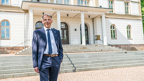 Professor Tolkmitt steht freundlich lächelnd vor dem Hauptgebäude der Hochschule Mittweida. Er trägt einen blauen Anzug, ein weißes Hemd und eine blaue Krawatte.