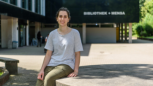 Das Fotos zeigt eine lächelnde junge Frau, die Studentin Ane Zuniga Gurruchaga, auf einem großen Tisch aus Beton sitzen. Im Hintergrund ist ein Gebäude zu erkennen mit dem Schriftzug "Bibliothek + Mensa".