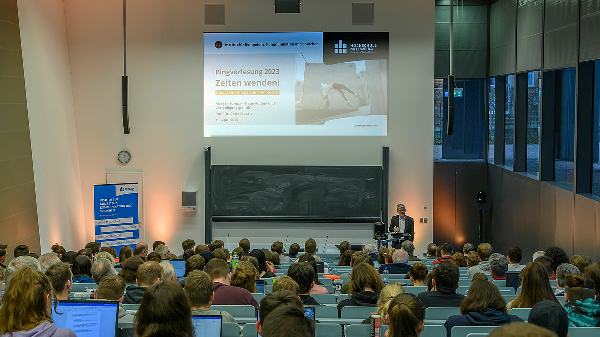 Blick von hinten über das Publikum im Hörsaal. Am Redner unten steht eine männliche Person, die zum Publikum spricht. Auf dem über ihr projizierten Beamerbild ist links neben der Grafik der Veranstaltungsreihe unter anderem zu lesen: „Ringvorlesung 2023. Zeiten wenden! ... Krieg in Europa – Neue Außen- und Verteidigungspolitik. Prof. Dr. Carlo Masala“.