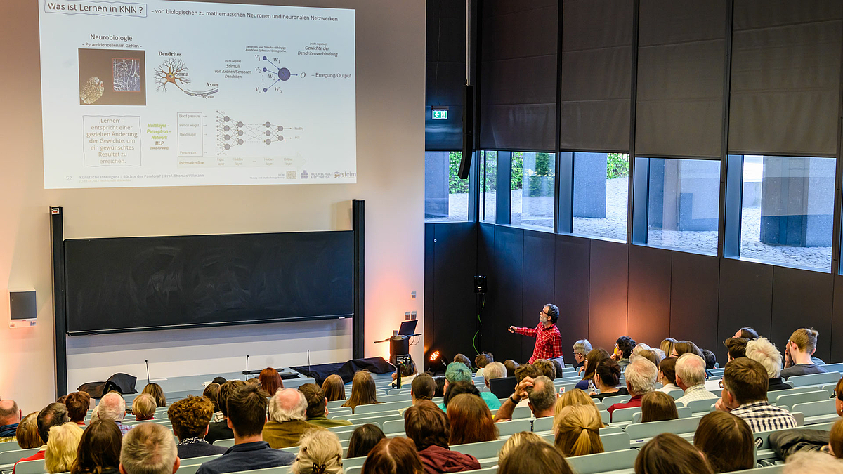 Blick von hinten in den Hörsaal über die Köpfe des Publikums auf den männlichen Dozenten. Schräg über ihm ist eine Folie projiziert mit der Überschrift: „Was ist Lernen in Künstlichen Neuronalen Netzwerken?“ Die Folie zeigt unter anderem mathematische Formeln.