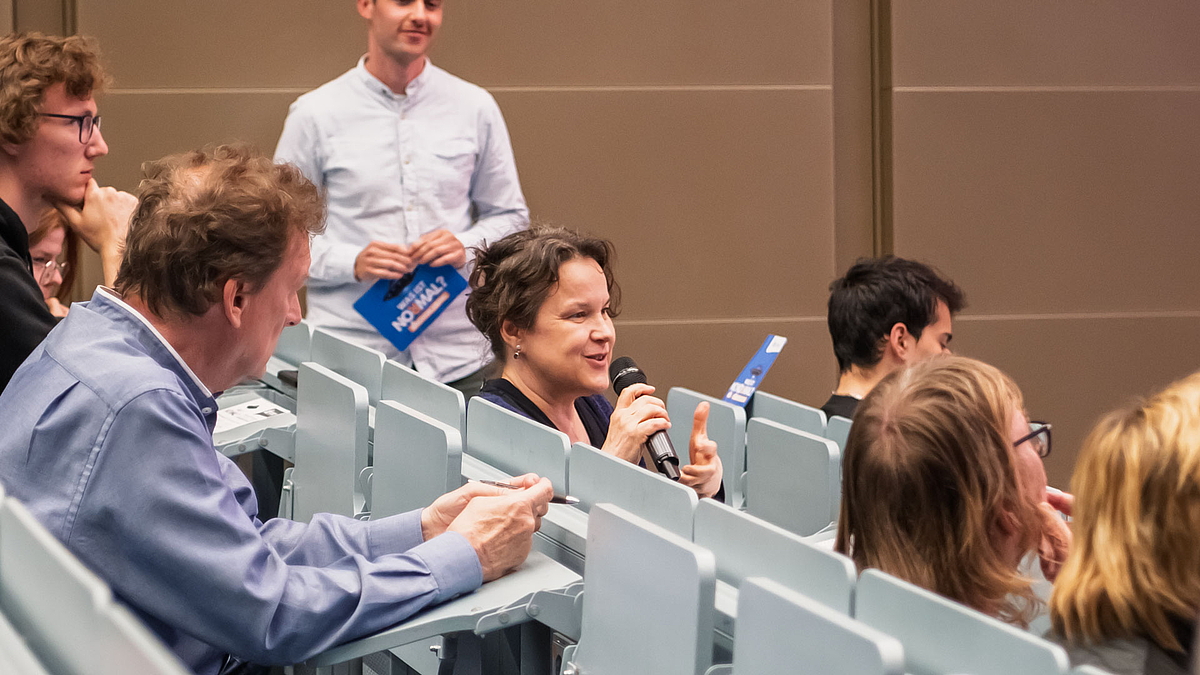 Eine Frau in einer der Bankreihen spricht in ein Handmikrofon- Um sie herum sitzen weitere Personen. Im Hintergrund steht ein Moderator.