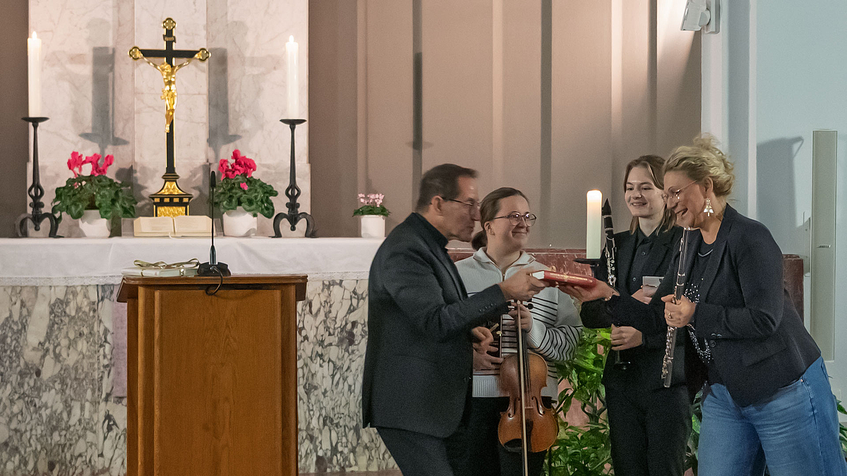 Alle vier Musiker:innen vor dem Altar. Der Musiker übergibt ein Geschenk.