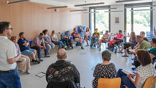 Eine Gruppe von rund 20 Menschen sitzt in einem Seminarraum in einem Kreis, die meisten auf Stühlen, zwei in einem Rollstuhl. Eine männliche Person steht am linken Bildrand der Gruppe zugewandt.