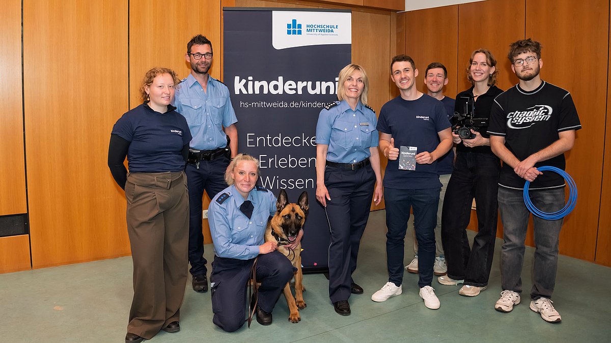 Ein Hund und acht Personen, darunter zwei Polizistinnen und ein Polizist, vor einem Rollup mit Kinderuni-Logo und dem Text „Entdecken, Erleben, Wissen“.