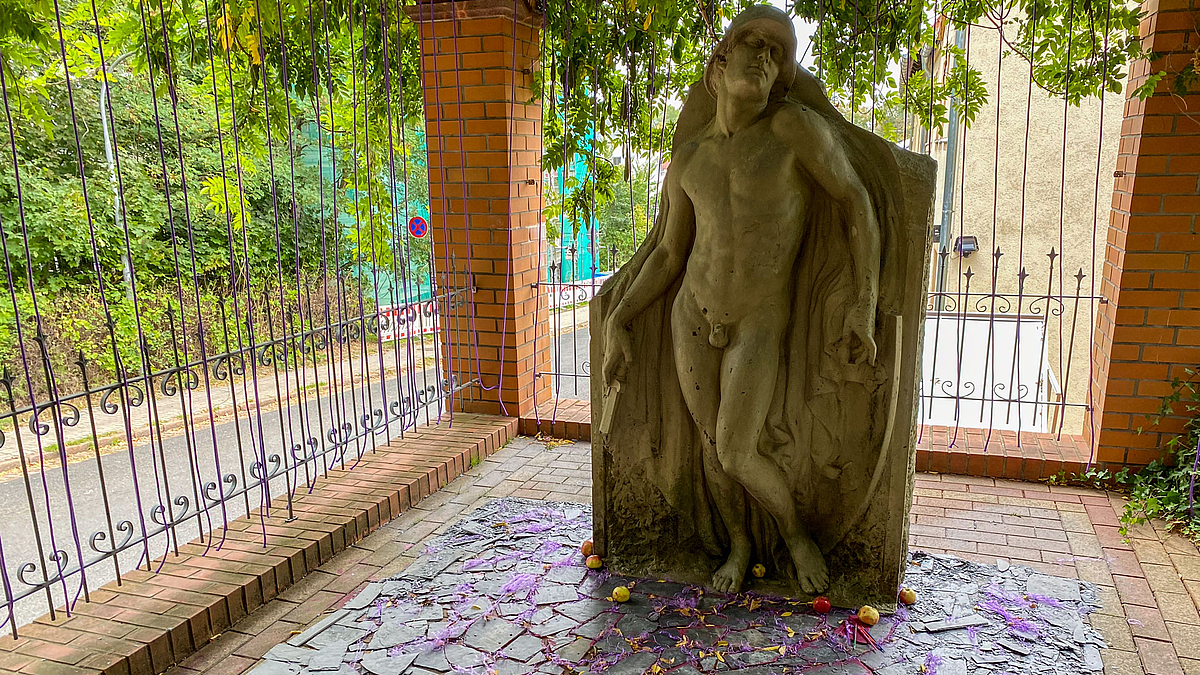 Historische Skulptur eines Soldaten am Ende einer Pergola. Am oberen Rand hängen zahlreiche lilafarbene Seile heran. Um das Denkmal herum sind Dachsschiefer, lilane Fäden und Äpfel gelegt.