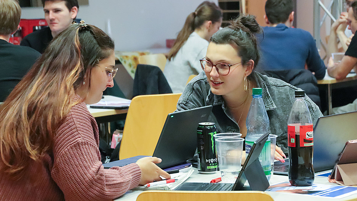 Zwei junge weibliche Personen an einem Tisch mit Laptops, Bechern, Colaflaschen und Energydrinks in Dosen. Die Frau rechts wendet sich der linken interessiert zu. Im Hintergrund sind weitere Personen an Tischen zu sehen.  