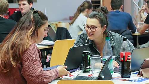 Zwei junge weibliche Personen an einem Tisch mit Laptops, Bechern, Colaflaschen und Energydrinks in Dosen. Die Frau rechts wendet sich der linken interessiert zu. Im Hintergrund sind weitere Personen an Tischen zu sehen.  