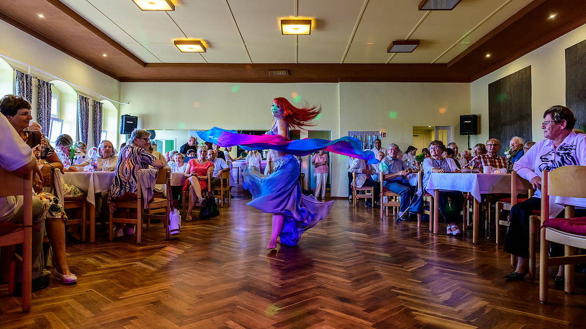 Mitten im Saal auf der freien Fläche zwischen den besetzten Tischen ist eine Bauchtänzerin beim Tanz zu sehen. Ihre roten Haare und ihr blaues Kleid wirbeln umher.